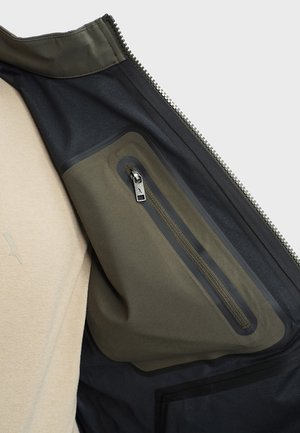 Inner jacket pocket with olive green waterproof zipper on dark fabric, visible next to beige shirt fabric background.