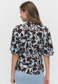 Korte mouwen blouse in lichtblauw met een contrasterend bruin bloemenpatroon. Zachte stof, losse pasvorm en een sluitingdetails aan de achterkant.