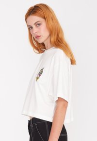 Camiseta blanca recortada hecha de algodón suave, con un diseño bordado colorido en el lado izquierdo del pecho. De corte holgado y mangas cortas.