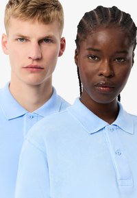 Magliette polo azzurre chiare con una texture a coste, dotate di un colletto classico e una patta con due bottoni, indossate da due persone posizionate vicino.
