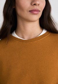 Sudadera color naranja quemado con cuello redondo, presenta una textura suave y sutiles motas multicolores, superpuesta sobre una camiseta blanca.