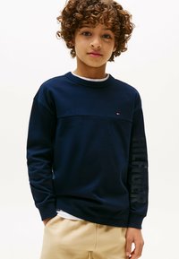 Felpa a maniche lunghe blu navy in cotone. Presenta un design orizzontale con un logo sulla manica e uno scollo a girocollo. Indossata sopra una camicia bianca.