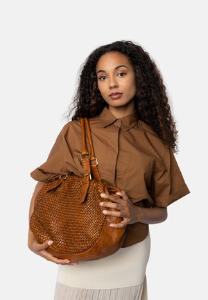 Giovane donna con capelli ricci tiene in mano una grande borsa in pelle intrecciata marrone, indossando una camicia marrone fluida e una gonna chiara su uno sfondo bianco.