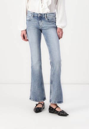 Kvinde iført lyseblå vidde jeans, en hvid langærmet bluse og sorte spidse flats med sølvnitter.