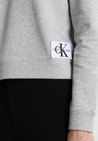 Sudadera gris con puños y dobladillo acanalados. Presenta un parche blanco con "CALVIN KLEIN JEANS" y un logo "CK" en negro. Textura de tela suave.