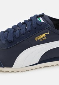 Mörkblå tygskor med vita sidostreck, guld "Puma Roma"-bokstäver och strukturerade gummisulor med ett zigzag-mönster.