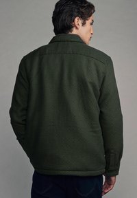Grüne texturierte Jacke mit einem Kragen-Halsabschluss, knopfbarem Bündchen und sichtbaren Nahtdetails; getragen von einer Person, die mit dem Rücken steht.