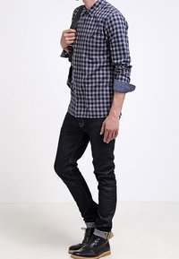 Zwart-witte geblokte shirt met opgerolde mouwen, gedragen met donkere jeans en zwarte laarzen met een gele accenten. Model houdt een tas vast.