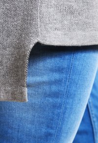 Šedý pletený svetr s žebrovanými lemy, mírně delší vzadu, umístěný nad modrými džínovými kalhotami, ukazující hladkou texturu.