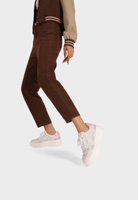 Sneakers bianchi con accenti rosa, pannelli testurizzati, punta rotonda e suola spessa; indossati con jeans marroni a vita alta e gamba affusolata e una giacca bomber multicolore.