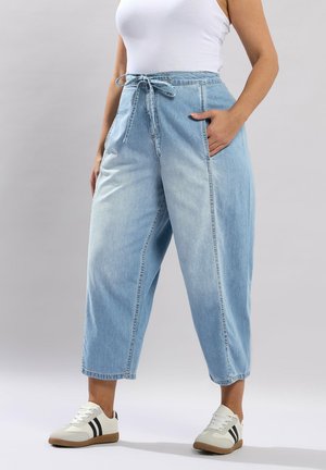 Kvinde iført lyseblå højtaljede jeans med vide ben og bælte, hvid tanktop samt hvide sneakers med sorte striber, stående.