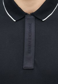 Κοντινό πλάνο του γιακα του μαύρου πόλο πουκαμίσου με άσπρη επένδυση και κάθετη μαύρη υφασμάτινη λωρίδα με ανάγλυφο το λογότυπο "ARMANI EXCHANGE."