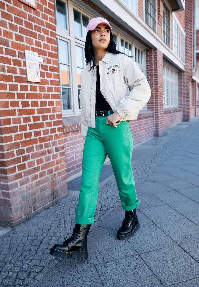 Beige Bomberjacke mit Logo, schwarzes Shirt, hellgrüne Jeans und schwarze Plateaustiefel, vor einer Backsteinmauer und Fenstern.