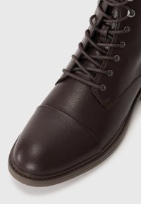 Botas de cuero marrón con cordones, superficie texturizada, punta redondeada y costuras más claras en contraste a lo largo de la suela.