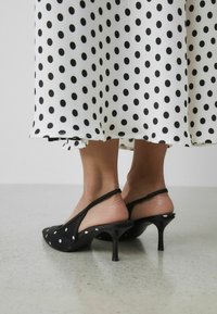 Escarpins à talons slingback noirs à pois avec un bout pointu, un petit talon et un tissu texturé ; dotés d'une lanière autour du talon et d'une semelle lisse.