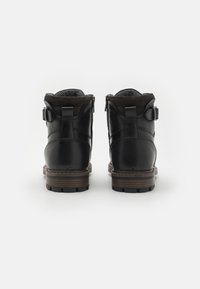 Botas de tobillo de cuero negro con un cuello de gamuza marrón texturizada, cierre de cremallera y correa con hebilla; suela de goma resistente con tacos prominentes.