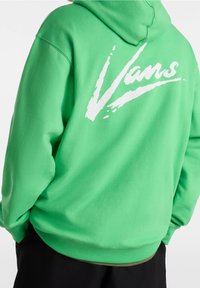 Osoba ubrana w jaskrawozieloną bluzę z kapturem z białym logo "Vans" na plecach, w połączeniu z czarnymi spodniami, na jednolitym białym tle.