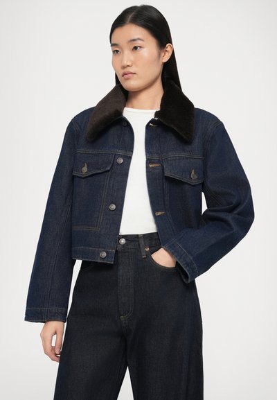 Tmavomodrá džínsová bunda s hnedým kožúškovým golierom, so zapínaním na gombíky, náprsnými vreckami a skráteným strihom. Nosená na bielom topánku.
