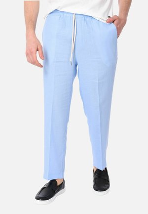 Uomo che indossa pantaloni azzurri con coulisse e scarpe nere slip-on, in piedi con una mano in tasca contro uno sfondo semplice.