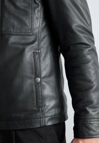 Gros plan sur la manche et le côté d'une veste en cuir noire avec un détail de poche à bouton-pression sur une personne portant un pantalon foncé.