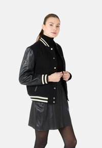 Zwarte varsityjack met leren mouwen, gestreepte geribde kraag en manchetten, met een witte nummer 73 accent. Gecombineerd met een zwarte leren rok.