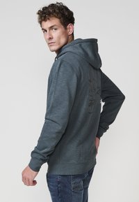 Szürke kapucnis pulóver textúrált felülettel és kontrasztos cérnával készült koponya mintával a hátsó részén; húzózsinóros kapucnival és bordázott gallérral rendelkezik.