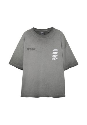 T-shirt gris oversize avec le texte "TRUE SELF" sur la poitrine gauche et quatre graphiques d'yeux blancs disposés en ligne verticale sur la poitrine droite.