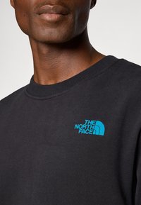 Close-up van een persoon die een zwart ronde hals sweatshirt draagt met een klein blauw "The North Face" logo op de borst.