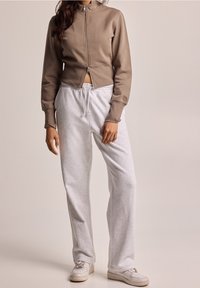 Felpa marrone con zip, tagliata, e colletto alto, abbinata a pantaloni sportivi grigio chiaro con cintura elastica e lacci.