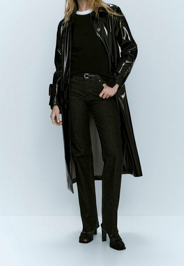 Personne portant un long manteau noir brillant, un pull noir, un pantalon noir et des chaussures noires à talons, debout devant un fond clair uni.