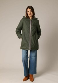 Veste imperméable verte avec capuche, dotée d'une fermeture éclair à l'avant et de détails rayés. Assortie à un jean bleu à jambes larges et des bottes marron.