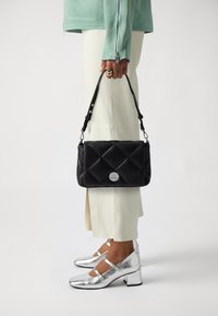 Svart quiltad handväska med en silverlogotyp, hålls av en person som bär silverfärgade klackskor och ljusa vida byxor.