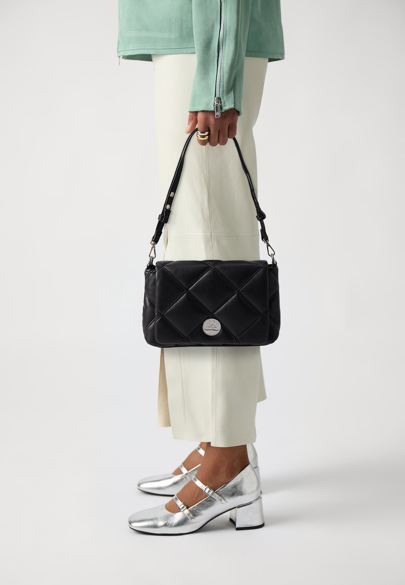 Schwarze gesteppte Handtasche mit einem silbernen Logo, gehalten von einer Person, die silberne Absatzschuhe und hellfarbige weit geschnittene Hosen trägt.