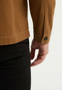 Gros plan sur la main droite d'une personne pendue à côté de son pantalon noir, portant une chemise à manches longues marron avec un poignet boutonné.