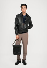 Zwarte leren bomberjack met twee voorzakken, een marineblauwe trui eronder, beige broeken en een zwarte bucket bag met een gestructureerde voorkant.