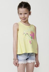 Rumena brezrokavnik s cvetličnim vzorcem, ki vključuje roza in oranžne akcentne barve, v kombinaciji z jeans kratkimi hlačami z čipkastim robom.