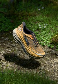 Scarpe da corsa arancioni e marroni con tomaia testurizzata, dettagli neri e suola ammortizzata, sospese sopra il terreno con erba e pietre sottostanti.