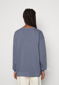 Överdimensionerad blå sweatshirt av bomullsblandning, med rundad halsringning, ribbade ärmslut och rak fåll; bärs med ljusa byxor.