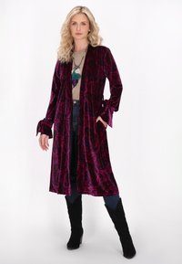 Karmesinrober mit langen Ärmeln, die ein Paisley-Muster aufweist, seitliche Taschen und Bindebänder an den Bündchen hat, über einem hellbraunen Oberteil getragen.