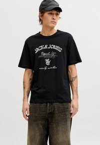 Zwart T-shirt met korte mouwen en witte grafische opdruk, voorzien van het "JACK & JONES" logo. Gecombineerd met vervaagde, wijde denim jeans.
