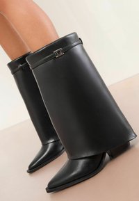 Fekete bőr bokacsizmák éles, hegyes orral és blokk sarokkal. Széles szegély díszíti fémból készült ékszerrel a tetején.