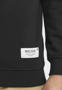 Černá mikina s žebrovanými manžetami a spodním lemem, na níž se nachází bílá značka s nápisem "INDICODE" a sloganem. Hladká textura, ležérní design.