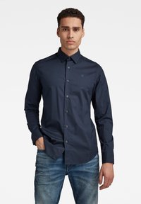 Camicia blu navy con bottoni, collo, maniche lunghe e un piccolo logo, abbinata a jeans blu sbiaditi, con una texture liscia.