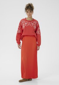 Sieviete ar cirtainiem matiem, ģērbusies sarkanā džemperī ar baltām ziedu kontūrām un garā pleitotā sarkanā svārkā, ar brūnajām sporta apaviem.