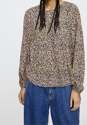 Personne portant un chemisier à manches longues à motifs floraux orange, blanc et noir, avec un jean bleu taille haute, debout devant un fond clair uni.