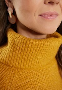 Maglione a collo alto in giallo senape con texture a costine. Presenta anche orecchini circolari e rettangolari, evidenziando un design moderno.