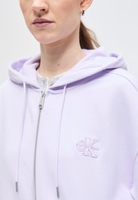 Άτομο που φοράει φούτερ με φερμουάρ σε ανοιχτό μωβ χρώμα με το λογότυπο της Calvin Klein στο στήθος και μικρό σκουλαρίκι κρίκο, ουδέτερο φόντο.