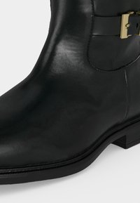 Bottine en cuir noir avec un petit talon et un détail de boucle dorée sur le côté.