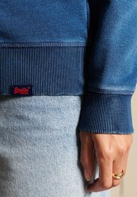 Niebieska bluza z dzianinowymi mankietami i dołem. Charakteryzuje się teksturowanym materiałem oraz małą czerwoną naszywką z logo. Nosi się ją na jasne dżinsy.