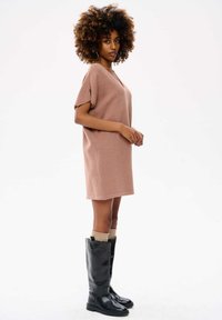 Blade różowy, oversize'owy sweter dress z dekoltem w kształcie litery V, w długości powyżej kolan, w połączeniu z wysokimi, czarnymi skórzanymi botkami i beżowymi skarpetkami.